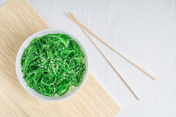 Wall Mural - Top view of chuka wakame seaweed vegetarian salad decorated with sesame seeds served for dinner in white bowl with chopsticks on bamboo mat on white wooden background. Horizontal orientation image