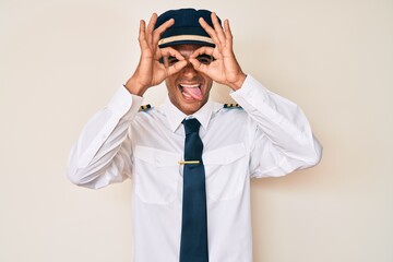 Sticker - Young hispanic man wearing airplane pilot uniform doing ok gesture like binoculars sticking tongue out, eyes looking through fingers. crazy expression.