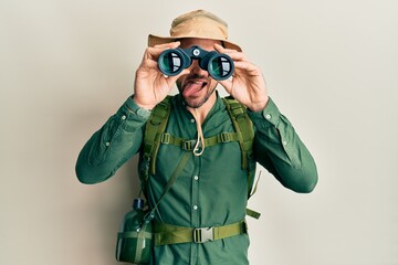 Poster - Handsome man with beard wearing explorer hat looking through binoculars sticking tongue out happy with funny expression.