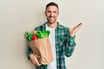 Poster - Handsome man with beard holding paper bag with groceries smiling cheerful presenting and pointing with palm of hand looking at the camera.