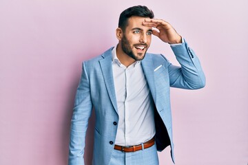 Young hispanic businessman wearing business jacket very happy and smiling looking far away with hand over head. searching concept.