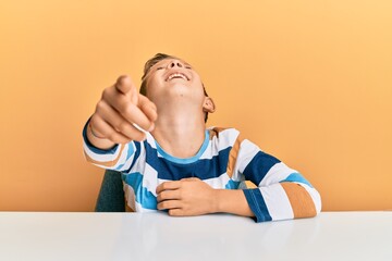 Adorable caucasian kid wearing casual clothes sitting on the table laughing at you, pointing finger to the camera with hand over body, shame expression