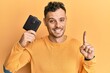 © Krakenimages.com - Young hispanic man holding leather wallet smiling with an idea or question pointing finger with happy face, number one