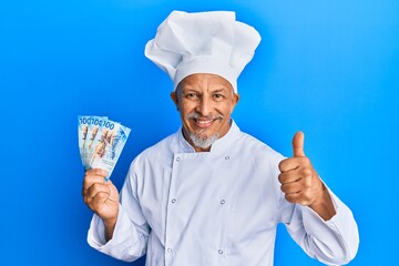 Canvas Print - Middle age grey-haired man wearing professional cook uniform holding swiss franc banknotes smiling happy and positive, thumb up doing excellent and approval sign