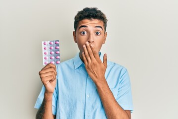 Sticker - Young handsome african american man holding pills covering mouth with hand, shocked and afraid for mistake. surprised expression