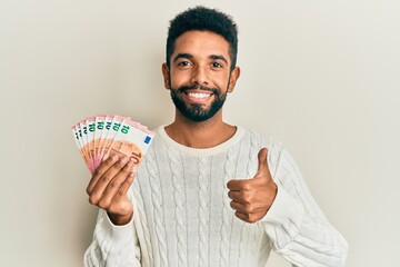 Wall Mural - Handsome hispanic man with beard holding bunch of 10 euro banknotes smiling happy and positive, thumb up doing excellent and approval sign