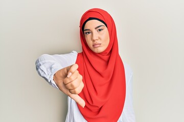 Wall Mural - Young beautiful hispanic girl wearing traditional islamic hijab scarf looking unhappy and angry showing rejection and negative with thumbs down gesture. bad expression.