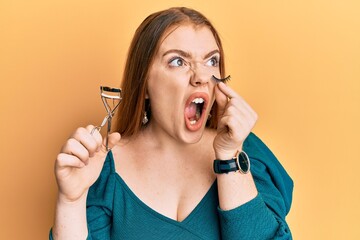 Canvas Print - Young beautiful redhead woman holding eyelash curler and fake lashes angry and mad screaming frustrated and furious, shouting with anger. rage and aggressive concept.