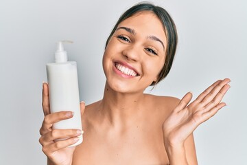 Young brunette woman holding cosmetic moisturizer facial cream celebrating achievement with happy smile and winner expression with raised hand