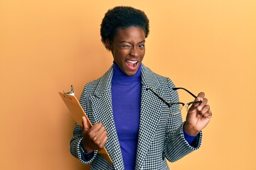 Poster - Young african american girl holding clipboard and glasses winking looking at the camera with sexy expression, cheerful and happy face.