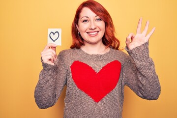 Poster - Young beautiful redhead woman holding reminder with heart over isolated yellow background doing ok sign with fingers, smiling friendly gesturing excellent symbol