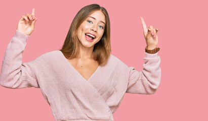 Beautiful blonde woman wearing casual winter pink sweater smiling amazed and surprised and pointing up with fingers and raised arms.