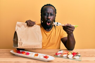 Poster - Handsome young black man eating take away sushi sitting on the table afraid and shocked with surprise and amazed expression, fear and excited face.