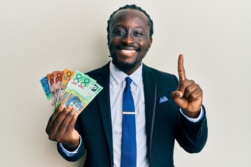 Sticker - Handsome young black man wearing business suit and tie holding australian dollars smiling with an idea or question pointing finger with happy face, number one