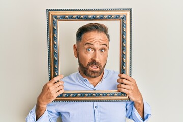 Poster - Handsome middle age man holding blank empty banner in shock face, looking skeptical and sarcastic, surprised with open mouth