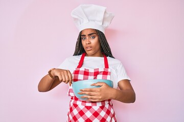 Canvas Print - Young african american woman with braids cooking using baker whisk clueless and confused expression. doubt concept.