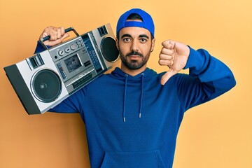 Canvas Print - Young hispanic man holding boombox, listening to music with angry face, negative sign showing dislike with thumbs down, rejection concept