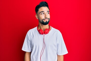 Wall Mural - Young man with beard listening to music using headphones smiling looking to the side and staring away thinking.