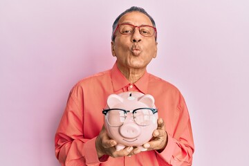 Poster - Middle age indian man holding piggy bank with glasses looking at the camera blowing a kiss being lovely and sexy. love expression.