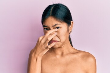 Canvas Print - Young chinese woman standing topless showing skin smelling something stinky and disgusting, intolerable smell, holding breath with fingers on nose. bad smell