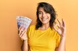 Young hispanic woman holding 20 polish zloty banknotes doing ok sign with fingers, smiling friendly gesturing excellent symbol
