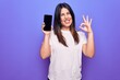 Young beautiful brunette woman holding smartphone showing screen over purple background doing ok sign with fingers, smiling friendly gesturing excellent symbol