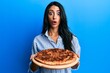 Beautiful hispanic woman holding tasty pepperoni pizza afraid and shocked with surprise and amazed expression, fear and excited face.