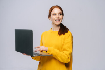 Charming happy young business woman or student holding laptop computer and looking away on isolated gray background. Pretty lady model with red hair emotionally showing facial expressions, copy space.