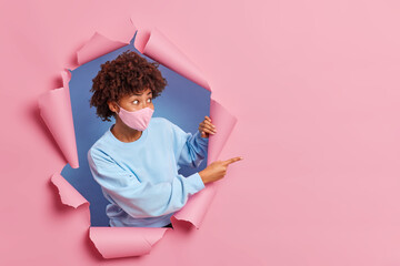 Wall Mural - Health care coronavirus and pandemic concept. Afro American woman in protective face mask shows announcement or promotion points away on blank empty space breaks through paper pink background.