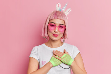 Portrait of good looking Asian woman with trendy hairstyle wears unicorn headband heart shaped sunglasses and t shirt makes thankful gesture isolated over pink background. Thank you very much