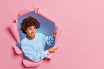 Canvas Print - Studio shot of thoughtful curly haired Afro American woman in casual clothes indicates at copy space advertises something breaks through paper background demonstrates logo. Promotion concept