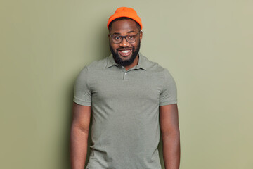 Wall Mural - Handsome bearded man with dark skin feels happy wears orange hat and t shirt isolated over khaki background. Afro American guy has cheerful expression smiles broadly poses indoor. Emotions concept