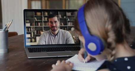 Wall Mural - On-line male tutor teaches little girl by videocall due covid-19, home-schooling concept. View over kid shoulder sit at table wear headphones listens teacher explanation writes down exercise solution