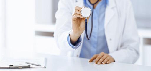 Unknown woman-doctor in blue blouse is holding stethoscope head while sitting at the desk in clinic, close-up. Physician ready to examine and help patient. Medicine concept