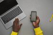 © Seventyfour - Minimal grey background of African-American male hands holding smartphone with blank screen over laptop while working at desk, top down view, copy space
