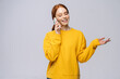 Smiling young woman wearing stylish yellow sweater talking by mobile phone on isolated white background. Pretty lady model with red hair emotionally showing facial expressions in studio, copy space.