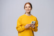 © dikushin - Happy smiling young woman holding mobile phone and looking at camera on isolated white background. Pretty redhead lady model emotionally showing facial expressions in studio, copy space.