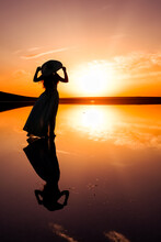 Silhouette Of A Girl In A Blue Dress On The Surface Of A Pink Lake. The Concept Of Freedom And Beauty. Sunset Photos