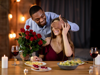 Wall Mural - Romantic Surprise. Happy black man covering girlfriend's eyes and giving roses bouquet