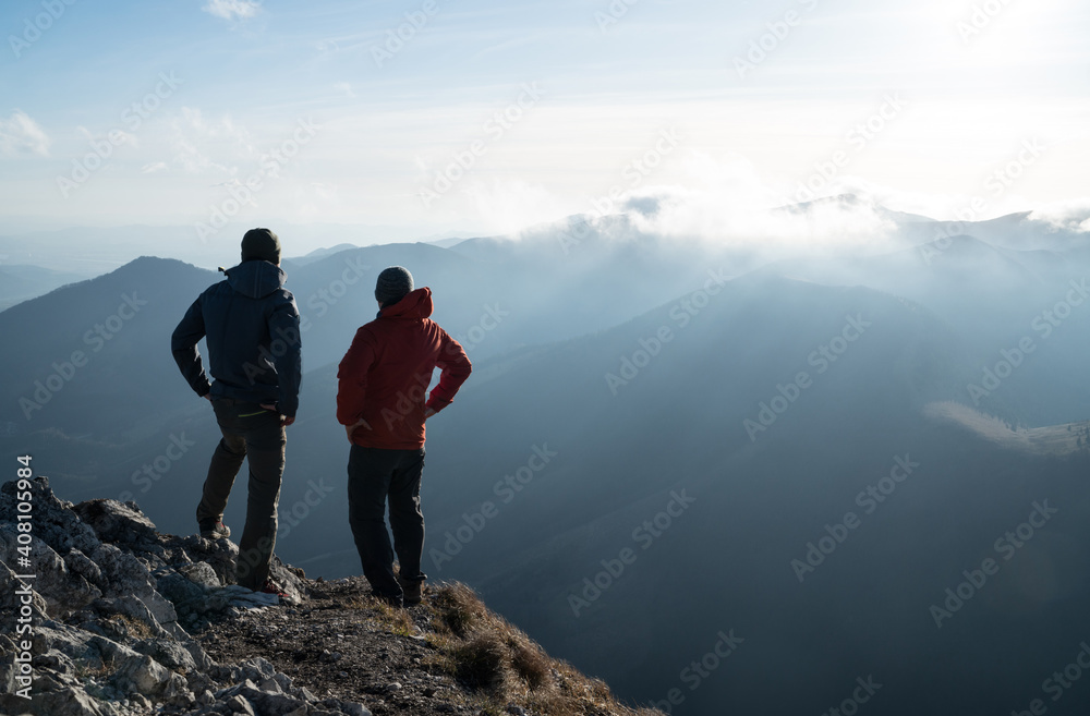 Two men standing standing with trekking poles on cliff edge and looking at sunset rays over the clouds. Successful summit concept image. - obrazy, fototapety, plakaty 