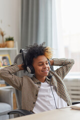 Wall Mural - Vertical portrait of smiling African-American boy wearing headset and relaxing in chair while using laptop at home, gamer or blogger concept