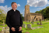 Fototapeta Na ścianę - A priest stands outside the historic St Etheldreda's Church in Wales. The older man is wearing a black shirt with a white clerical collar. There is a cemetery in front of the church.