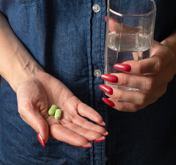 a woman in a denim shirt holds a glass of water in one hand and in the other hand two tablets on her palm. illness, headache, treatment.