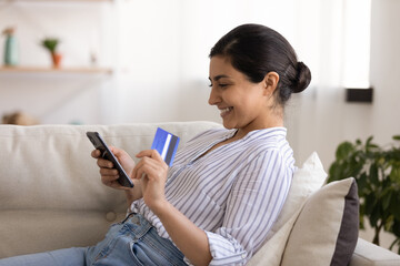 Young indian woman shopper sit on couch enjoy easy paying for goods online using bank card phone. Happy mixed race female traveler book tickets trip hotel at travel agency transport operator website