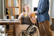 Young businesswoman in casualwear passing cup of tea to her disable colleague