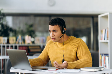 Confused male manager sits at the desk, using computer. Hispanic businessman have a problems with internet connection while online conference. Young adult employee worried about work issues