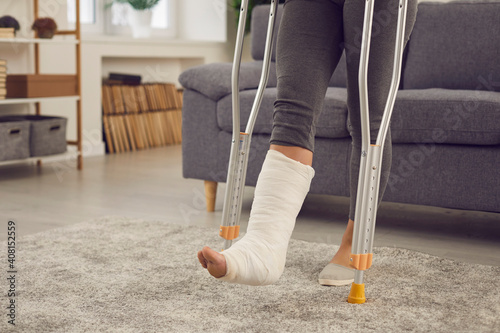 Concept of injury in domestic or car accident, rehabilitation, making progress and successful recovery. Young woman with broken leg in plaster cast stands up from couch and walks with crutches at home