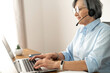Senior mature female gray-haired call center operator sitting at the desk in the home office, wearing a headset, answering the call, writing an email to the customer, typing on the laptop
