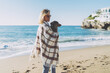 Charming blonde woman wearing blue jeans with a plaid on her shoulders standing with her dog in her arms on a blurred background of a sea shore. Student girl walking the beach side with her pup.
