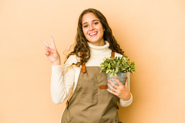 Wall Mural - Young caucasian gardener woman holding a plant isolated joyful and carefree showing a peace symbol with fingers.